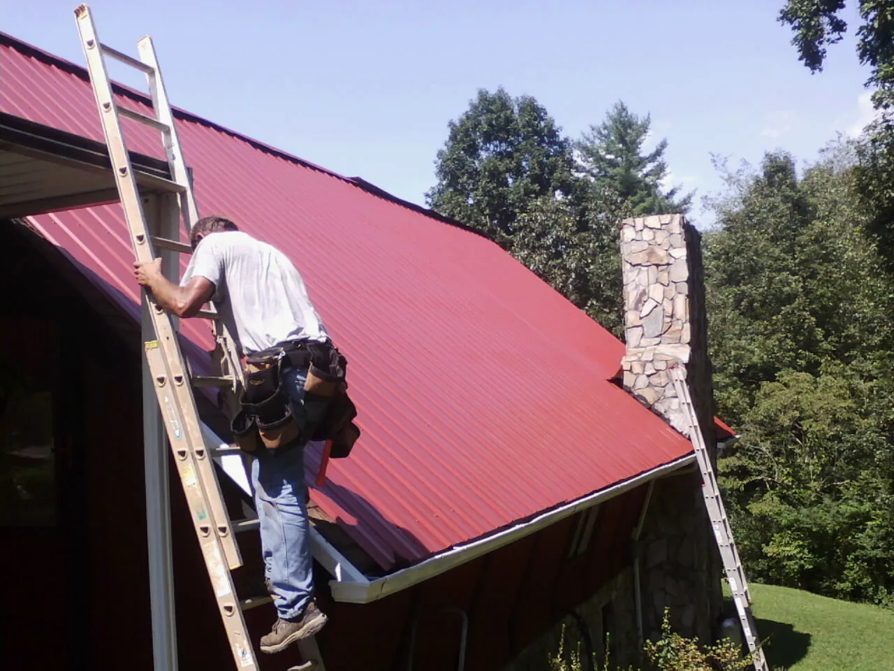 Roofer on a ladder beside a red metal roof and stone chimney — Soffit & Fascia Repair by Roofing Brothers in Kiln